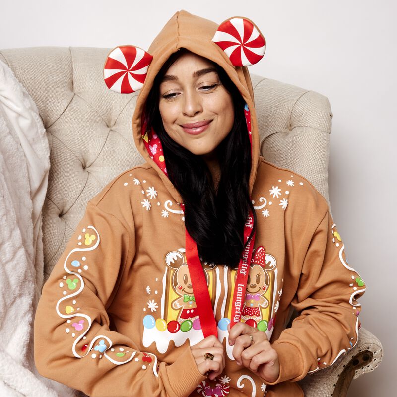 Woman sitting in a white chair wearing the Mickey and Friends Gingerbread House Unisex Hoodie with the hood up, showing off the peppermint candy Mickey Ears on the hood.